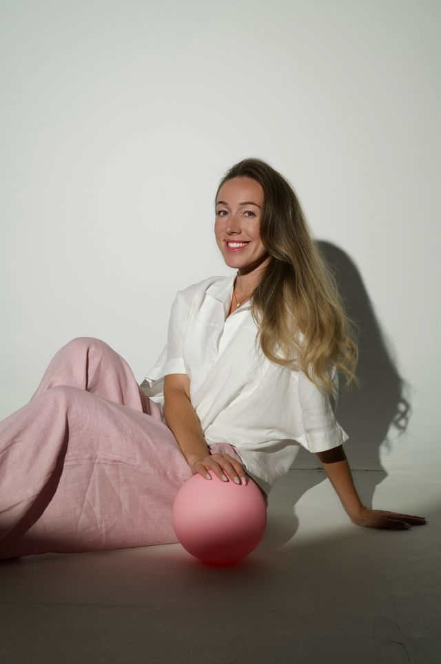 Chloe seated on the floor in white linen and blush trousers resting a hand on a pink Pilates ball
